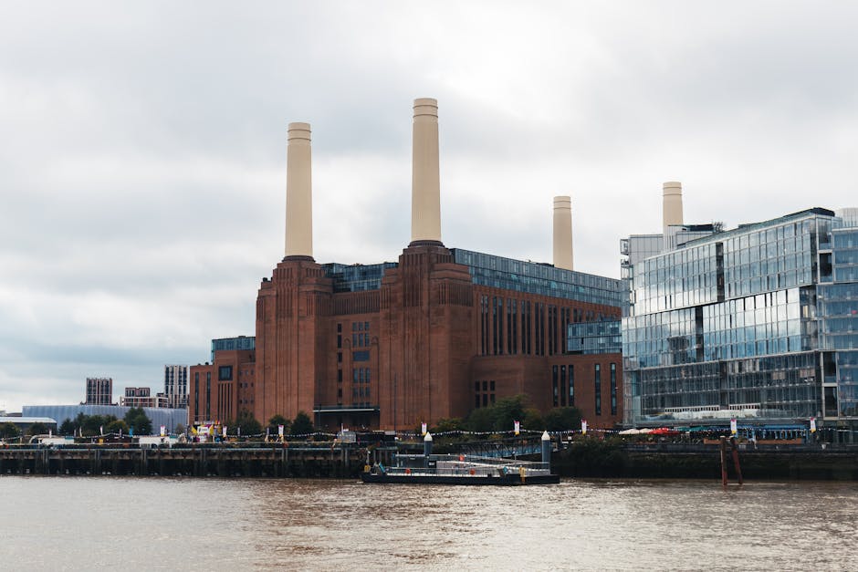 A large red brick industrial building with four tall, cream-colored chimneys situated along the riverbank under an overcast sky, with modern glass-fronted office buildings adjacent on the right. In the foreground, there is a quay with a docked boat, and the water appears brownish. Behind the building, a lamppost and some signage are visible, along with trees and distant structures. The scene depicts an urban riverside area likely involved in local transport or industrial activity. This image could be used to illustrate the environment around house removals or moving logistics in Battersea, with reference to the area's transport infrastructure. The inclusion of the building's structural details and the river setting provides context for the relevant location mentioned on the page about affordable removals from Battersea Park to Clapham Junction, referencing the neighborhood's blend of industrial heritage and modern development, relevant for services like those offered by Man With a Van Battersea.