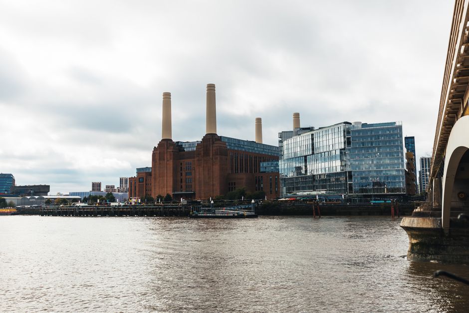 A view of a large industrial building with three tall smokestacks, situated along the riverbank in Battersea. To the right, part of a bridge with an arched structure extends over the water, which has