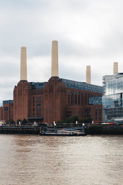A view of the iconic Battersea Power Station, a large brick building with four tall chimneys, situated alongside the River Thames on a cloudy day. The building features modern glass extensions on its sides, and the water in the foreground reflects the structure. In a house removals context, it is common for [COMPANY_NAME], such as Man With a Van Battersea, to assist with furniture transport and packing during relocations near the Battersea Power Station area, supporting the logistics involved in home relocation or moving services. This image highlights the importance of careful planning in loading and moving items from the property, with the power station serving as a recognizable landmark in the vicinity of London.
