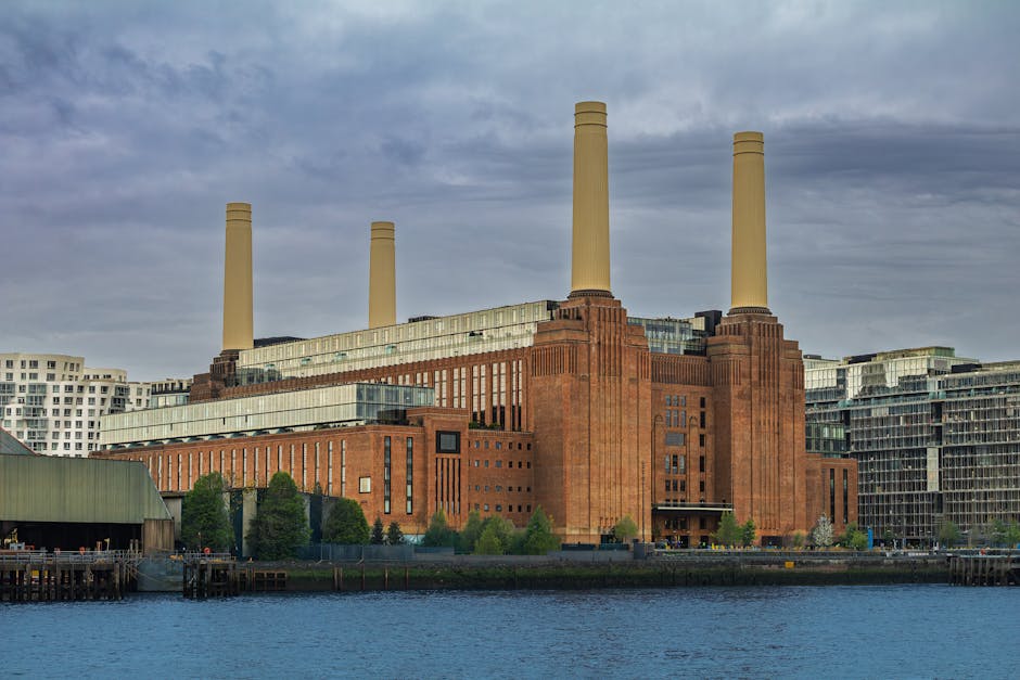 A large brick building with two tall white chimney stacks against a cloudy sky, serving as the iconic Battersea Power Station. In front of the building, there is a paved plaza with a series of illuminated metal sculptures depicting dancing human figures. The sculptures are mounted on black bases and arranged in a line that extends across the foreground. The scene is captured during daytime with natural lighting, and a few individuals are visible walking or sitting near the sculptures. To the sides of the power station, modern glass and steel structures are partially visible. The image emphasizes the scale of the historic power station, its brickwork, and the artistic outdoor installation, as part of a home relocation or moving process visual context for Man With a Van Battersea.